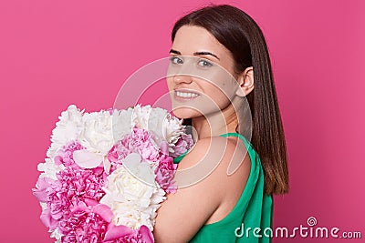 Profile of young brunette female with amazing bouquet of peonies, attractive woman wearing green sundress, has charming smile, Stock Photo