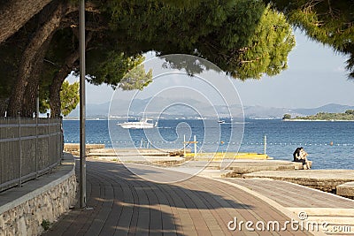 Promenade with pine trees by the sea and people and boats in the distance Editorial Stock Photo