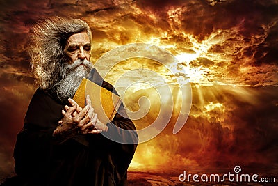 Prophet with Bible. Old Monk with Golden Book praying over Epic Landscape Background. Senior Bearded Man Worship in Black Cloak Stock Photo