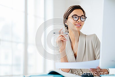 Prosperous businesswoman focused in monitor of computer, studies financial issues, holds document and pen, works in office, wears Stock Photo