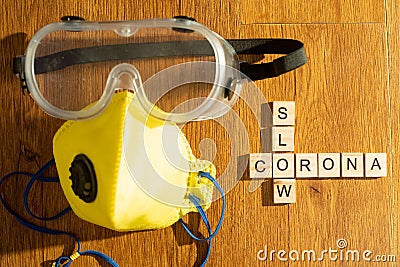 Protective eyewear and yellow mask with outlet valve with wooden blocks spelling out slow corona on a wood floor Stock Photo