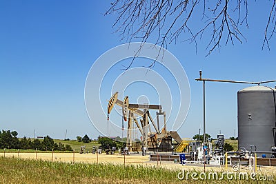 Two pump jacks at a pumping station with tanks and machinery with some farm buildings on the horizon under a blue sky with a few t Stock Photo