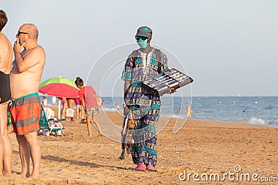 Punta Umbria, Huelva, Spain - August 7, 2020: African immigrant selling their wares on the beach wearing protective or medical Editorial Stock Photo