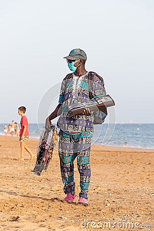 Punta Umbria, Huelva, Spain - August 7, 2020: African immigrant selling their wares on the beach wearing protective or medical Editorial Stock Photo