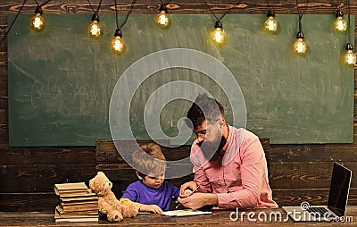 Pupil with teacher at school. Tutor helping kid to write letters in copybook. Man and boy sit at desk with laptop and Stock Photo