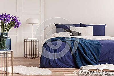 Purple flowers in blue glass vase on stylish table in white bedroom interior with comfortable bed Stock Photo