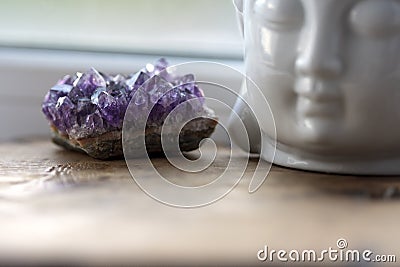 Purple and gemmy amethyst stone with White Buddha head on the windowsill background. Rough ametist crystals on home Stock Photo