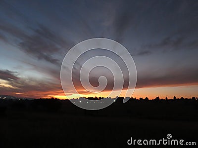 Purple, grey, blue and orange clouds form artistic dashes in the sky at sunset over silhouette of landscape Stock Photo