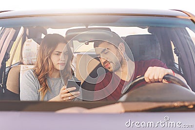 Puzzled female holds smart phone, uses online maps, shows to husband, try to find way, being lost in big city, drive car, have jou Stock Photo