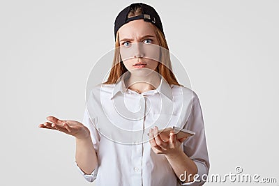 Puzzled female with stunned expresson, wears fashionable cap, shrugs shoulders in bewilderment, waits for call, holds modern smart Stock Photo