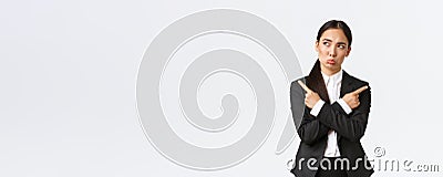 Puzzled pretty asian businesswoman in black suit troubled making choice, standing indecisive, pointing fingers sideways Stock Photo