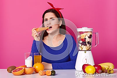 Puzzled woman keeps to healthy diet, bites fresh carrot, makes smoothie with food processor, wears red headband, blue sweater, Stock Photo