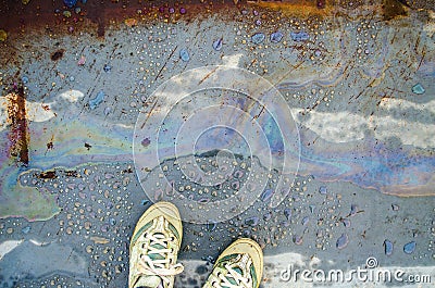 Rainbow chemical stains on the water in a puddle on road. Legs in sneakers. Stock Photo