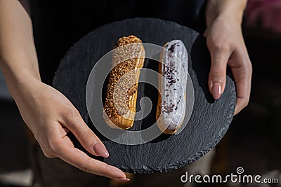 Raspberry and caramel eclair on round black rock stand in hands. Two eclairs with white and brown glaze in cafe. Cake Stock Photo