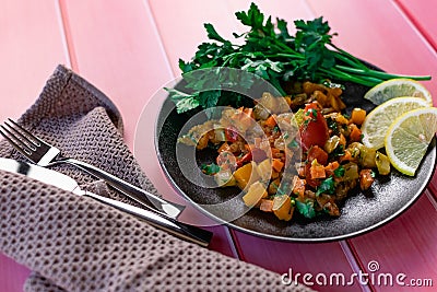 Ratatouille on a black plate, colored different vegetables in a stew, with fresh parsley leaves and lemon slices, on a pink wooden Stock Photo