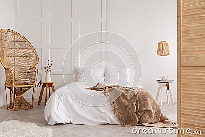 Rattan chair and wooden table next to bed with brown blanket in white bedroom interior. Real photo Stock Photo