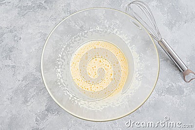 Raw beaten eggs with sugar in a glass bowl on a gray concrete background. Preparation of cream, scrambled eggs, French toasts, Stock Photo