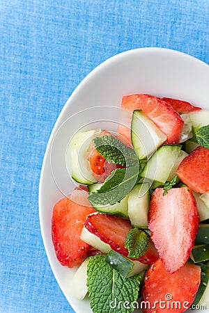 Raw fruit detox salad with cucumbers strawberries apples fresh mint in crystal bowl on blue table cloth background. Vegan Stock Photo