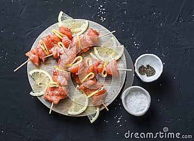 Raw ingredients for cooking salmon skewers on the grill. Raw salmon skewers, lemon, pepper, sea salt on a cutting board on dark ba Stock Photo