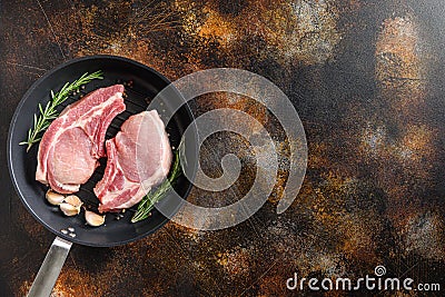 Raw Pork Loin chops on a bone in a black frying grill pan on a old rustic dark metall surface rosemary garlic peppercorns top view Stock Photo