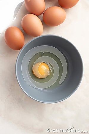 Raw uncooked egg in gray bowl and brown whole eggs on kitchen table, top view. Yellow egg yolk and liquid egg whites Stock Photo