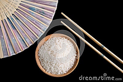 Raw white rice, hand fan and chopsticks on black background. Long uncooked rice in a wooden plate. Stock Photo