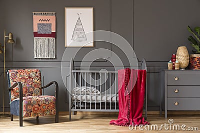 Real photo of a baby crib with a red blanket, an armchair, lamp Stock Photo