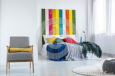 Real photo of a bright bedroom interior with white walls, comfy armchair and colorful blankets on a double bed Stock Photo