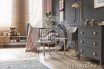 Real photo of a cot standing next to an armchair, lamp and cupboard in dark and classic baby room interior Stock Photo