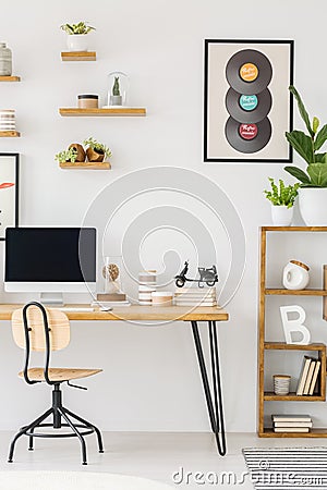 Real photo of a computer screen on a desk standing next to a chair and a shelf with ornaments and plants in bright work room Stock Photo