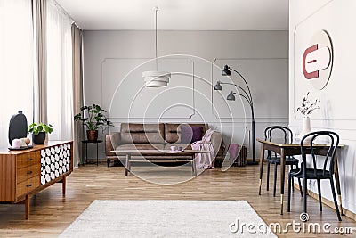 Real photo of light grey living room interior with window with curtains, leather couch, table with two chairs carpet on wooden flo Stock Photo