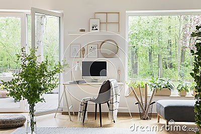 Real photo of white living room interior with big window, glass door, fresh plants, wooden desk with mockup computer and simple po Stock Photo