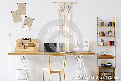 Wooden desk with a laptop, sewing machine, organizer and macrame o a wall next to a shelf. Empty screen, place you Stock Photo