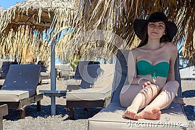 Realistic portrait of a young woman in a green swimsuit and hat, sitting on a chaise longue under a straw umbrella Stock Photo