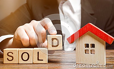 A realtor holds blocks with the word Sold near a miniature wooden house. Real estate market. Selling a house, apartment. Trade of Stock Photo