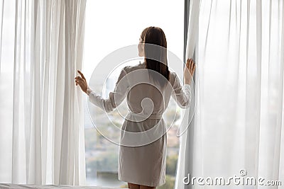 Rear back view young woman standing near opened curtains. Stock Photo