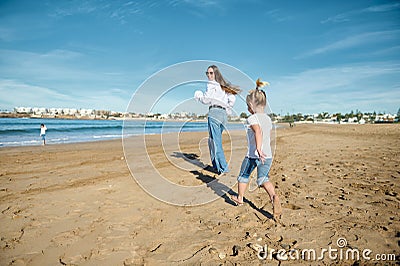 Rear view of a happy young mother and daughter smiling while running on the beach, leaving footsteps on the wet sand Stock Photo