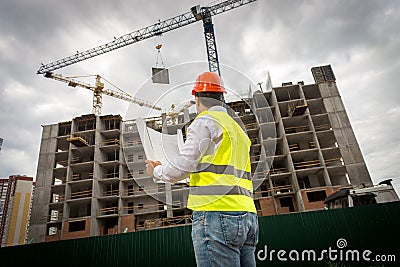 Rear view image of construction engineer in green safety vest and red hardhat controlling construction of new building Stock Photo