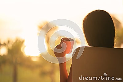 Rear view of woman with headphones and cup of coffee sitting on chair near window and looking on palm trees sea beach at sunrise. Stock Photo