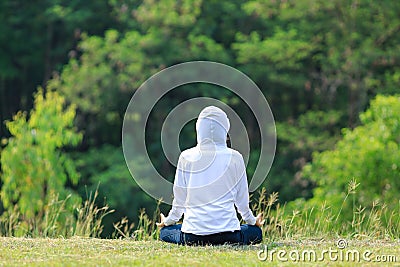 Rear view of woman in hoodie is relaxingly practicing meditation yoga in forest full of grass meadow in summer to attain happiness Stock Photo