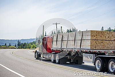 Red big rig semi truck transporting wood lumber on the flat bed semi trailer running on the turning road Stock Photo