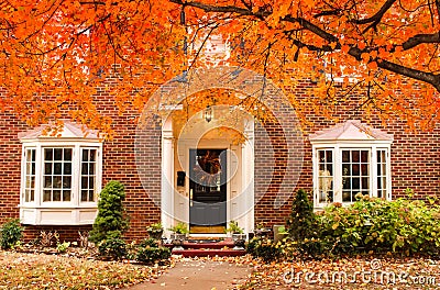 Red brick house entrance with seasonal wreath on door and porch and bay windows on autumn day with leaves on the ground and hydrag Stock Photo