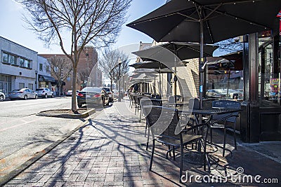 A red brick sidewalk dining area with black metal tables, chairs and umbrellas with cars parked along the street Editorial Stock Photo