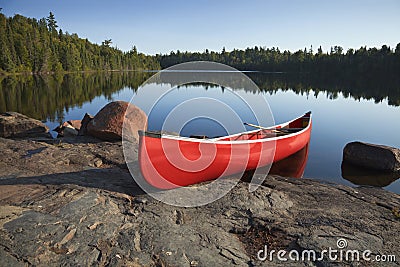 Red Canoe on Rocky Shore of Calm Lake with Pine Trees Stock Photo