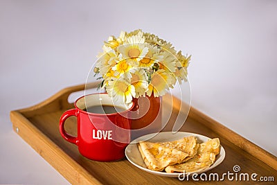 Red cup with the word `love` with hot coffee / tea, a bouquet of daisies and pancakes with honey on a wooden tray Stock Photo