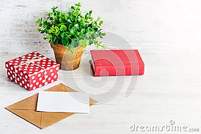 Red dotted gift box, empty card, kraft envelope, red book and a green flower in a rustic ceramic pot. White wooden background, cop Stock Photo