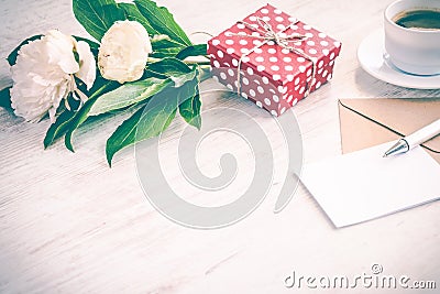 Red dotted gift box, kraft envelope and empty greeting card, coffee cup and peony flowers bouquet over white wood background. Copy Stock Photo