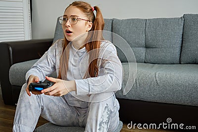 Red girl with tails in glasses sits on the floor and enthusiastically plays joystick. Stock Photo