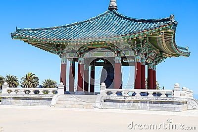 A red and green Korean Pavilion with massive bronze bell in the center with stone stairs on top of a hill with blue sky Stock Photo