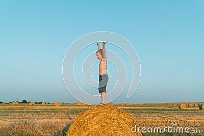 A red-haired boy stands with hands up on top of a straw bale on a wheat field Stock Photo